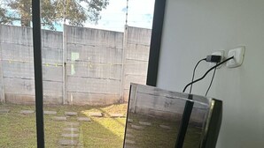 Ein Zimmer mit einer großen Schiebeglasdoor, die auf eine Terrasse mit Betonboden und einem Rasenbereich führt. Ein schwarzer Stuhl und ein kleiner Tisch mit einem Laptop stehen dort.