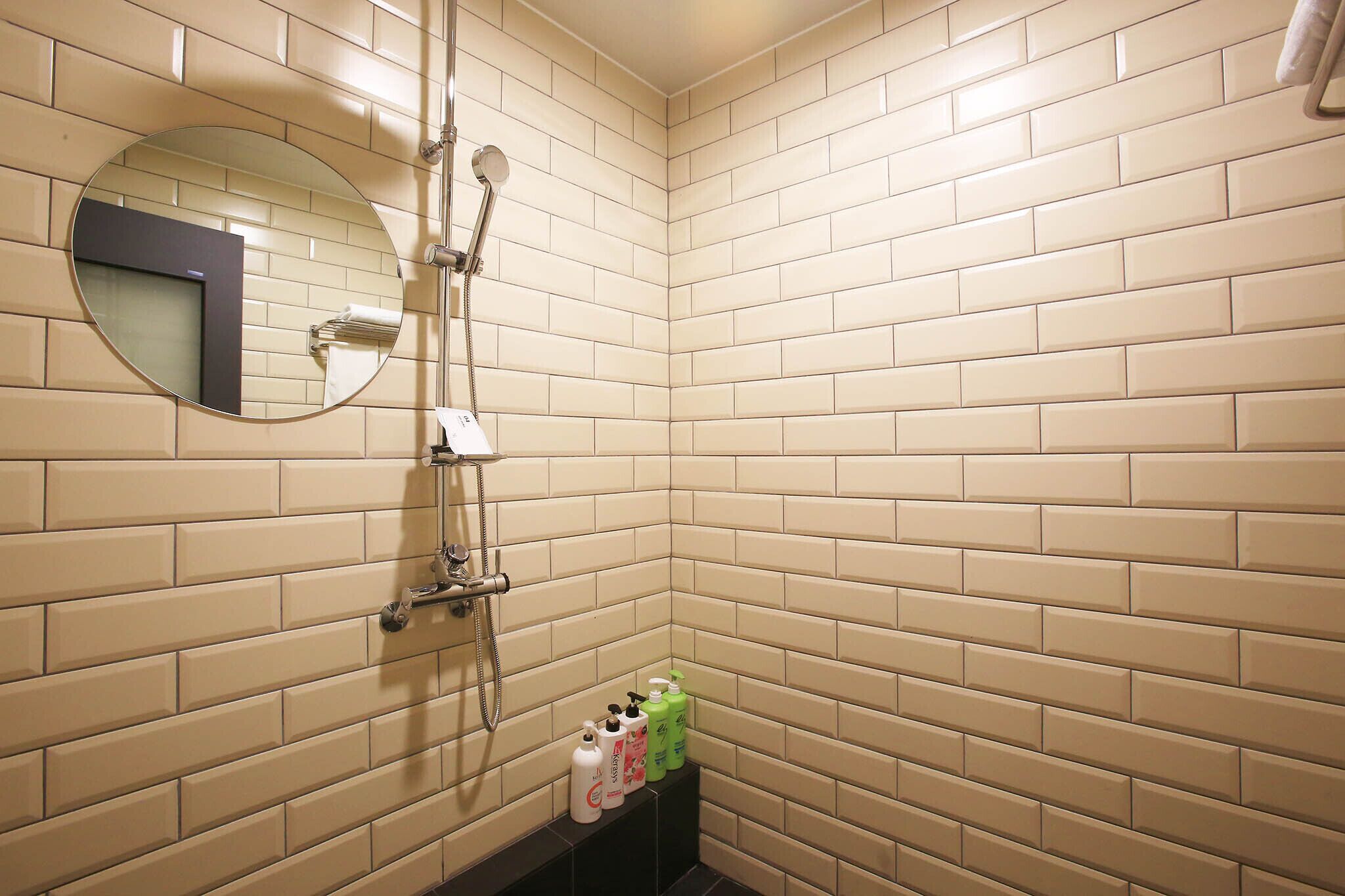 A corner bathroom with beige tiled walls, a round mirror, a showerhead, and a shelf with toiletries.