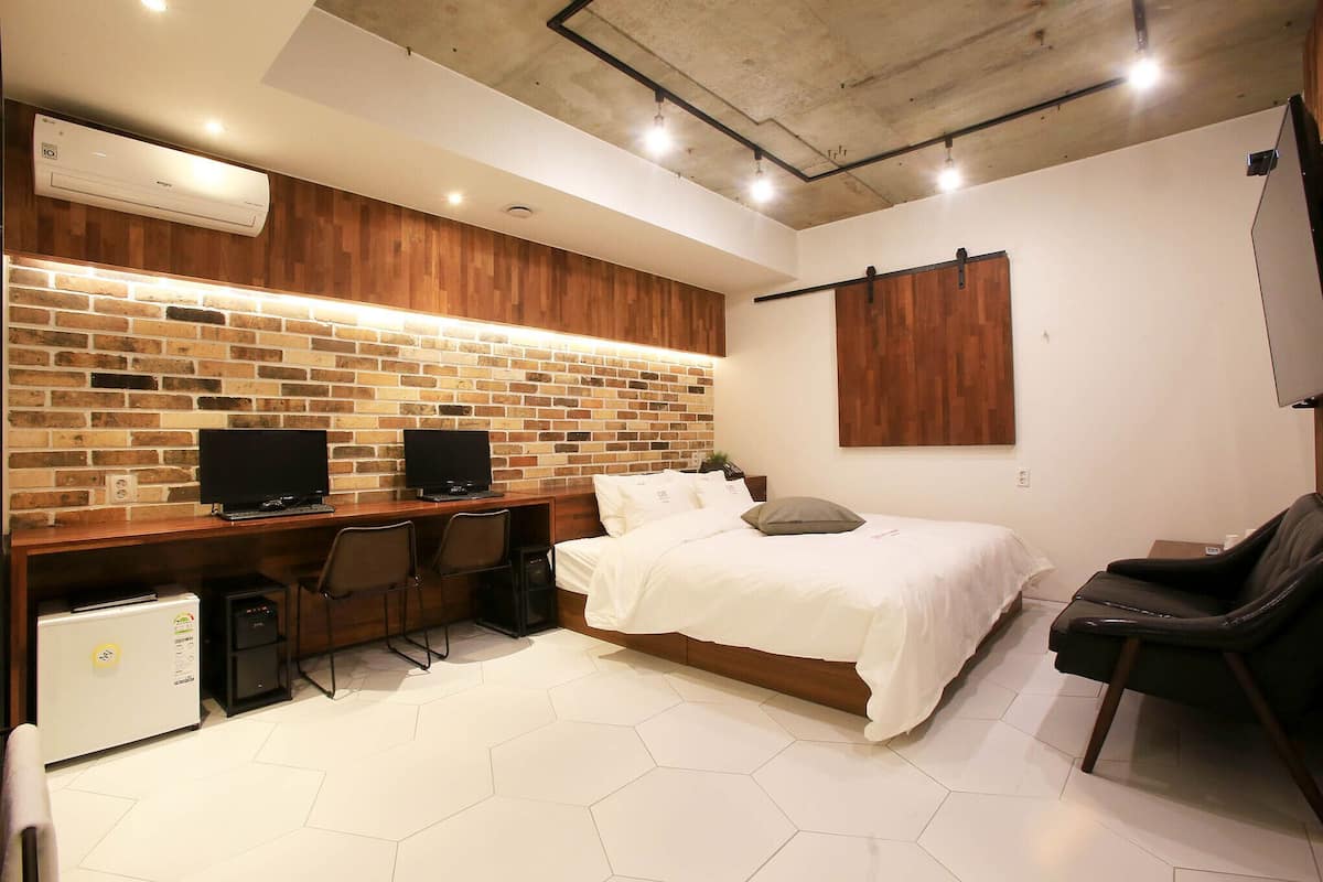 Ein modernes Hotelzimmer mit Backsteinwand, einem Bett, einem Schreibtisch mit zwei Monitoren, einem Kühlschrank und einem Sessel.