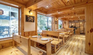 Un intérieur en bois, avec des bancs et des tables en bois, un coin cuisine et de grandes fenêtres donnant sur l’extérieur.