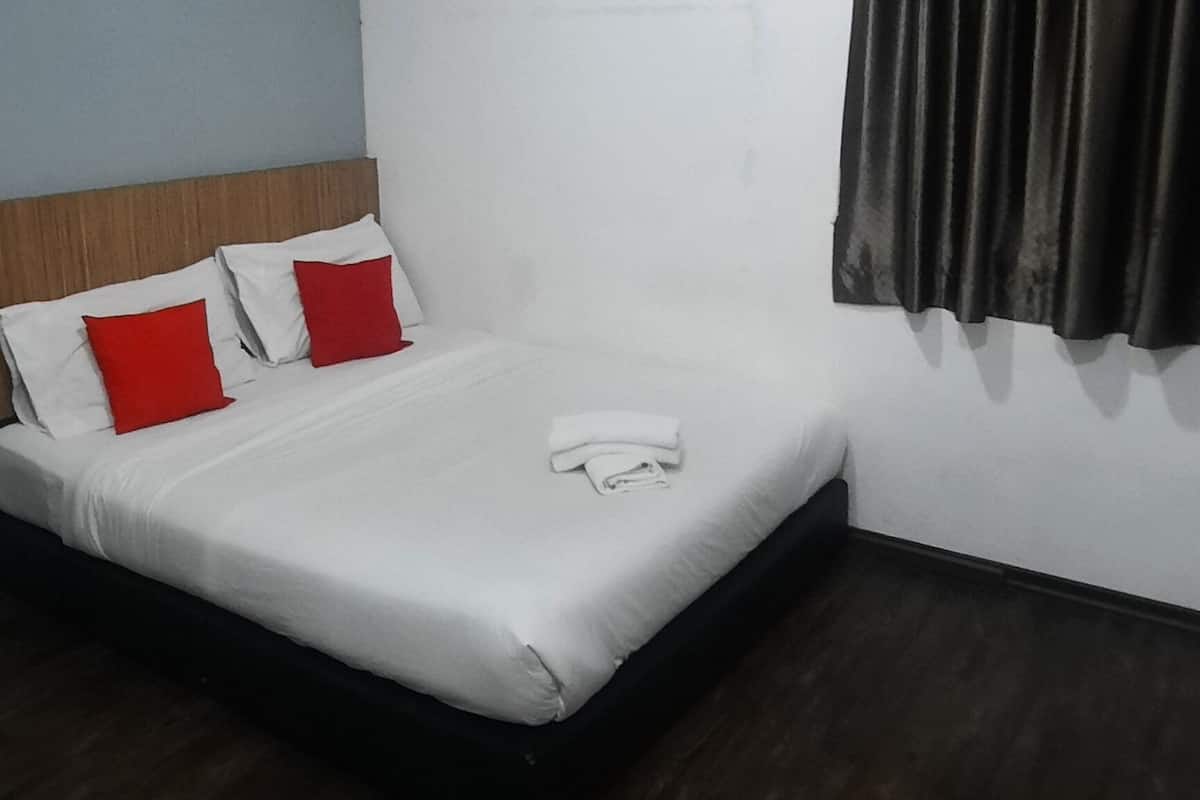 A hotel room with a bed, white walls, a wooden headboard, red pillows, a white blanket, and a dark curtain.