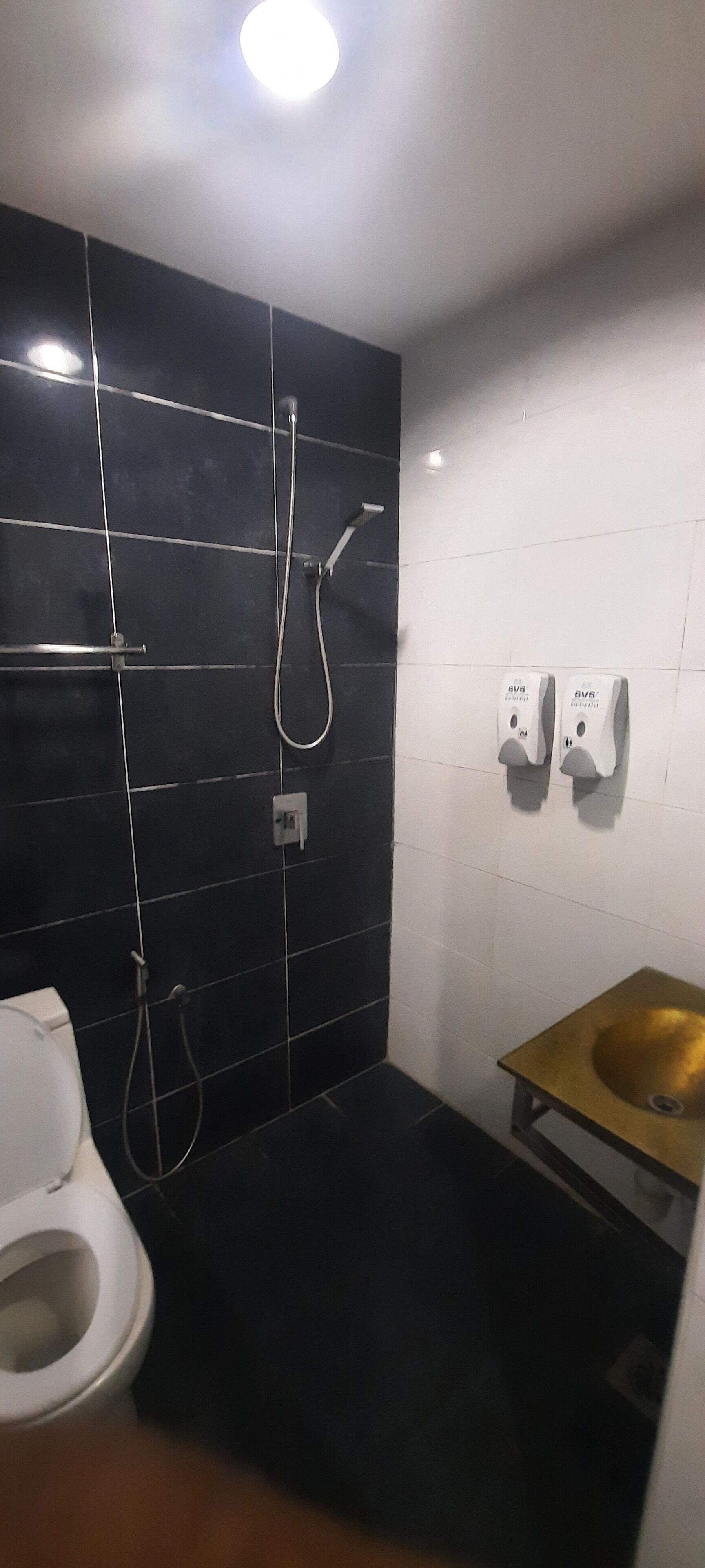A modern bathroom with a black tiled wall, a white toilet, a gold sink, and two hand soap dispensers.