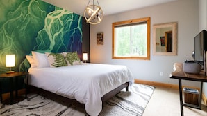 Une chambre avec un grand lit, un mur décoré d’une fresque abstraite aux tons verts, une fenêtre donnant sur des arbres et une table de chevet en bois avec une lampe.
