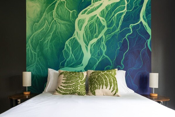 A bed with white linens, two pillows with a fern pattern, and a wall with a green abstract design.