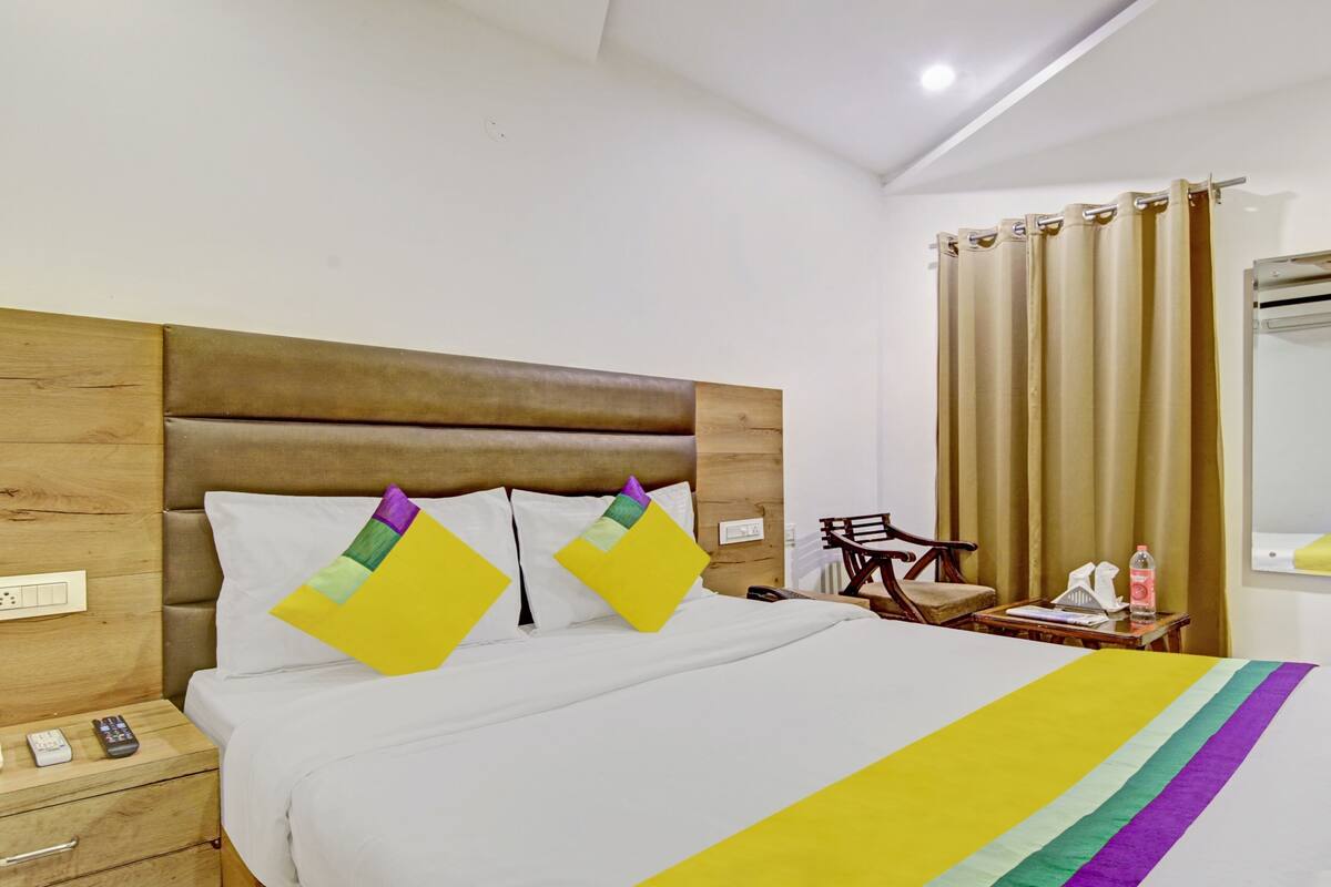 A hotel room with a wooden headboard, a bed with white bedding, a bedside table with a lamp, a chair, and a mirror.