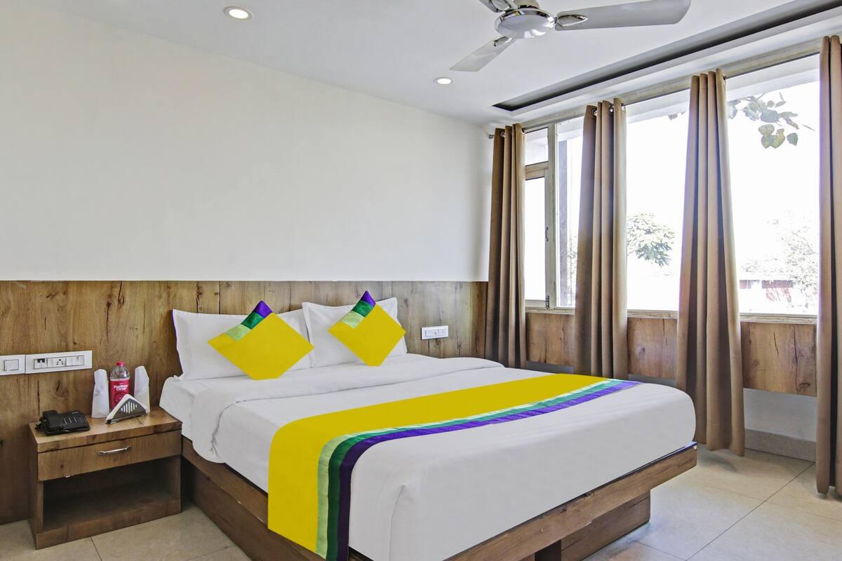A hotel room with a bed, wooden headboard, bedside table, and a window with curtains.