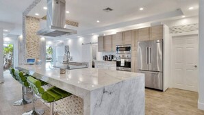 A modern kitchen with a marble island, stainless steel appliances, and a bar area with green barstools. - Lynora`s Luxe Estate - 3BR/3BA w/ Modern Design & Heated Pool (Fort Lauderdale)