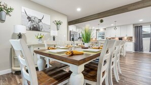 Un comedor con una mesa de madera dispuesta para seis personas, rodeada de sillas blancas. Se observa una cocina al fondo con gabinetes blancos y electrodomésticos de acero inoxidable.