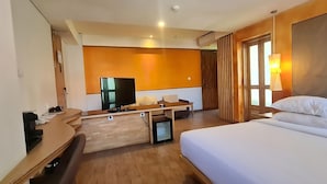 A hotel room with a bed, a desk with a flat-screen TV, a sofa, and a window with curtains. - Liberta Hotel Jimbaran (Kedonganan)