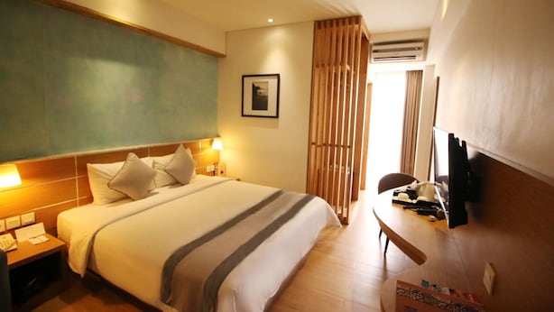 A hotel room with a large bed, bedside lamps, a desk with a chair, a television, and an air conditioning unit. - Liberta Hotel Jimbaran (Kedonganan)