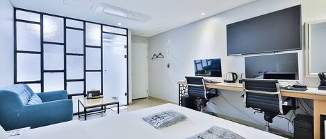 Habitación de hotel moderna con una cama grande, un escritorio con dos monitores, un sillón azul y vistas al mar a través de amplias ventanas.
