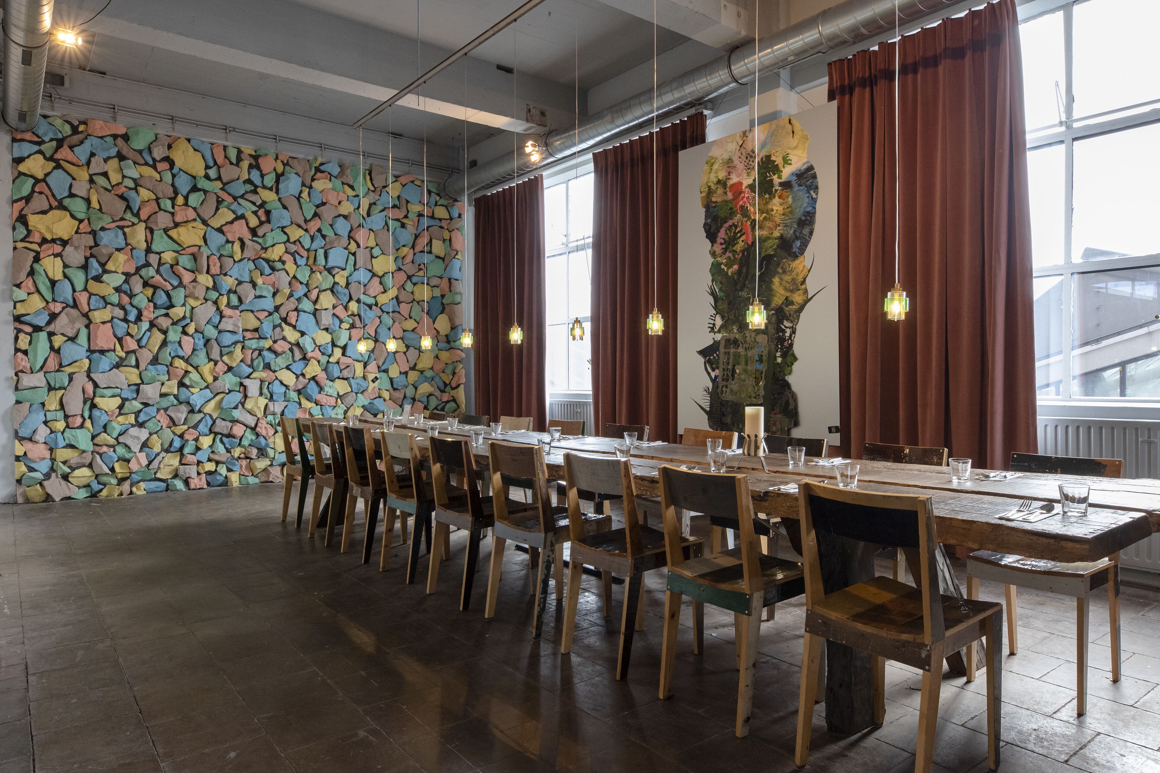 Une longue table à manger entourée de chaises, un mur décoré d’une mosaïque colorée et un grand tableau abstrait.