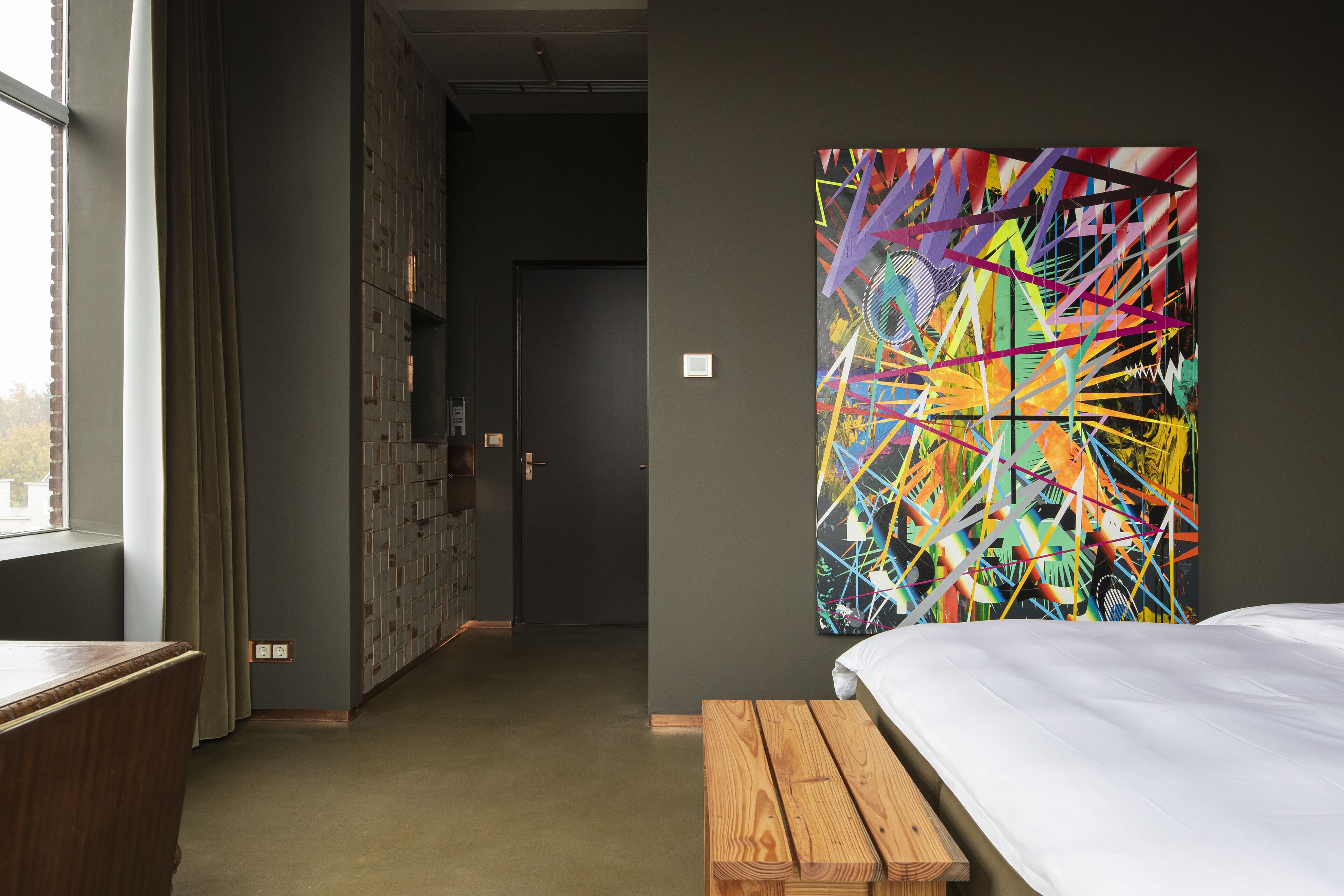 A hotel room with a large abstract painting on the wall, a wooden bench, and a bed with white bedding.