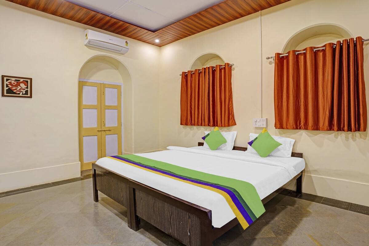 Habitación de hotel con cama, techo de madera, unidad de aire acondicionado y dos ventanas con cortinas.