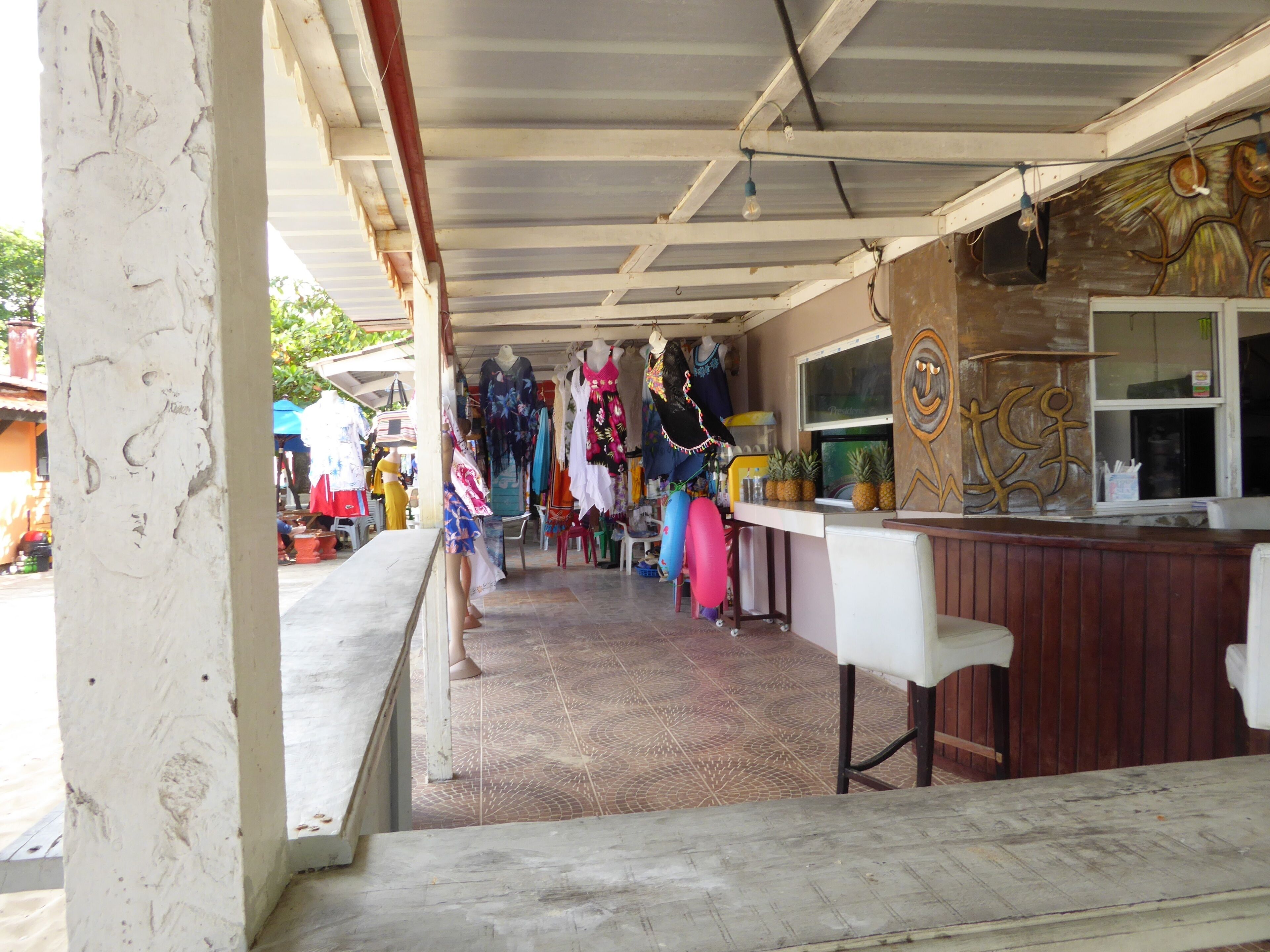 A covered outdoor area with a bar counter, various clothing items displayed on mannequins, and a tiled floor.