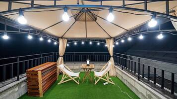A covered outdoor seating area with a table and two chairs, surrounded by a railing and artificial grass.