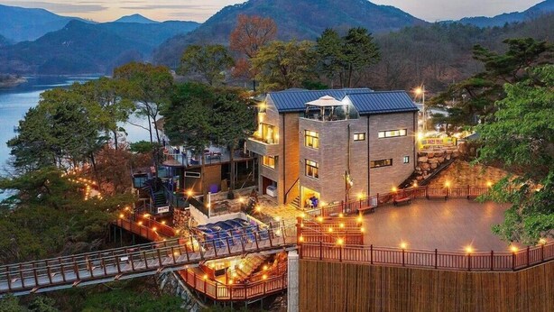 Un immeuble à plusieurs étages, doté d’un toit plat, entouré d’une terrasse en bois et d’une balustrade, et offrant une vue sur une étendue d’eau et des montagnes.
