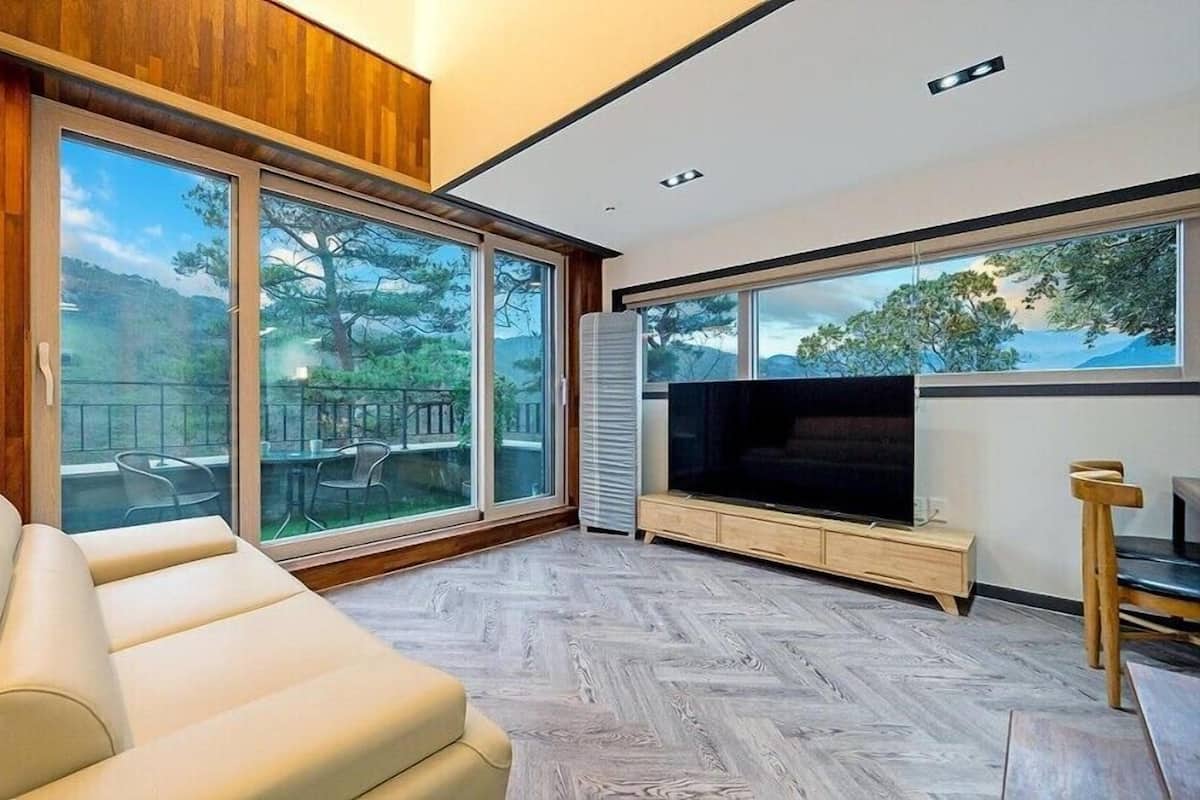 A modern living room with a sofa, a flat-screen TV, and large windows offering a view of the outdoors.