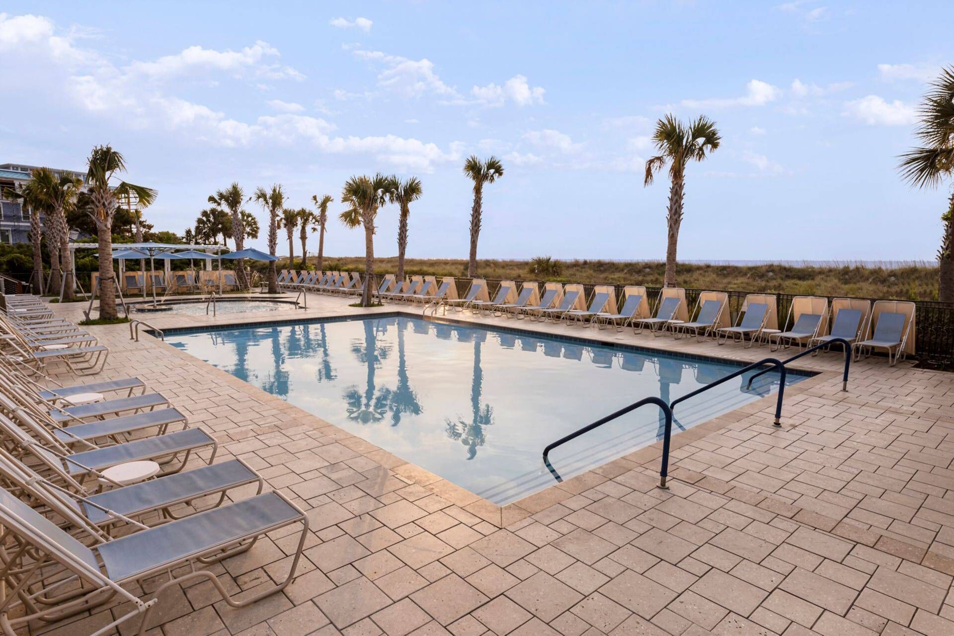 Un espace aménagé au bord de la piscine, avec des chaises longues et des palmiers, un ciel dégagé et une vue sur la plage.