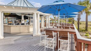 A covered outdoor seating area with wooden tables and chairs, a bar with a sign reading "STAY SALTY", and a gazebo with a blue umbrella. - Courtyard View - Marriott's SurfWatch - Upscale Resort On The Beach! (Hilton Head Island)