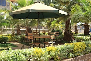 Een terras buiten met een tafel en stoelen onder een grote parasol, omgeven door palmbomen en verzorgde struiken.