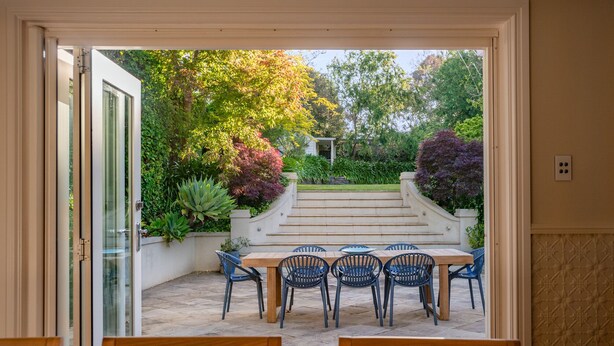 A patio with a table and chairs, surrounded by a garden with various plants and a staircase leading to a higher level. - Forest View House (Stirling)