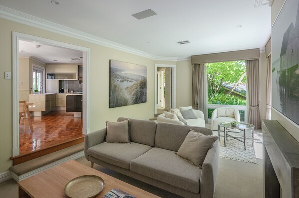 A modern living room with a sofa, a coffee table, and a TV. There is a kitchen visible through an open door.