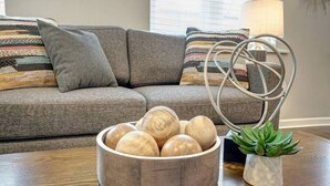 Un bol en bois contenant des boules en bois, posé sur une table en bois, un canapé gris avec des coussins à rayures et une petite plante en pot.