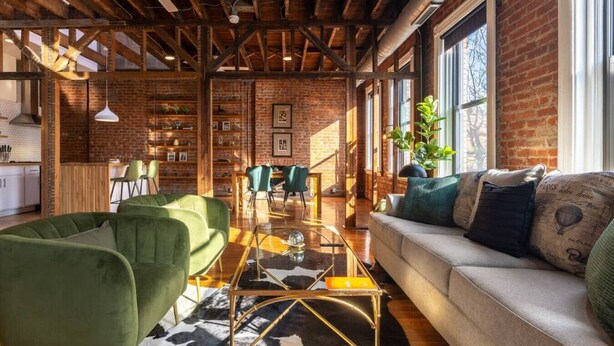 Um amplo loft com vigas de madeira expostas, parede de tijolos e janelas grandes. Há um sofá verde, um sofá branco e uma área de refeições com cadeiras.