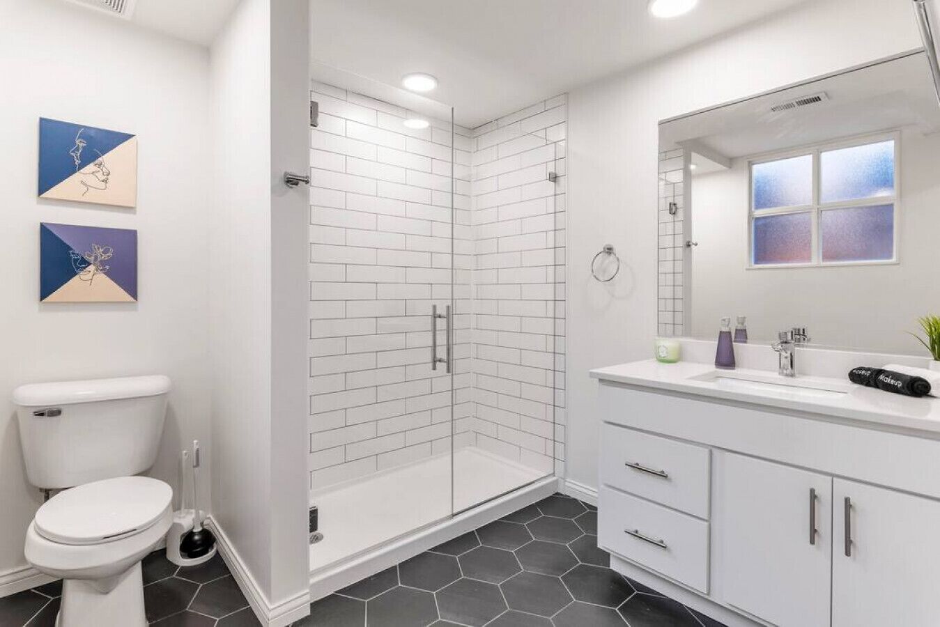 Une salle de bain moderne avec une douche à parois de verre, des carreaux blancs, des toilettes, un lavabo avec un miroir et deux œuvres d’art accrochées au mur.