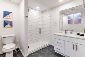 A modern bathroom with a glass-enclosed shower, white tiles, a toilet, a sink with a mirror, and two wall-mounted artworks. - Deluxe 3BR Loft Walkable to Everything Downtown (Cincinnati)