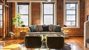 Un salon chaleureux avec un canapé et deux fauteuils, de grandes fenêtres, un mur en briques et un plancher en bois.
