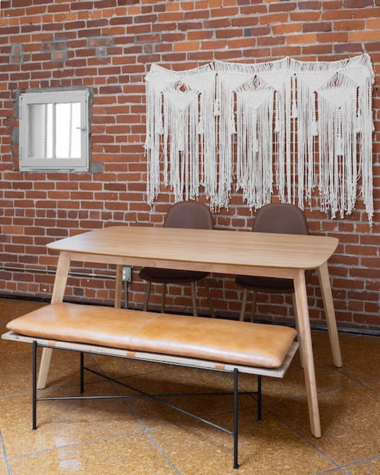 Een eetgedeelte met een houten tafel en bank, een bakstenen muur en een macramé wanddecoratie.