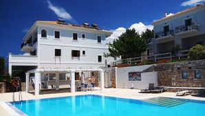 Un espace piscine avec une eau d’un bleu limpide, des bâtiments blancs dotés de balcons et un mur de pierre orné d’une fresque.