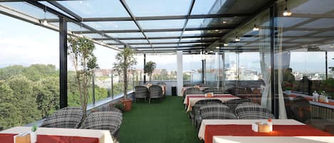 Un área de comedor al aire libre con mesas y sillas, piso de césped artificial verde y vistas a la ciudad y montañas.
