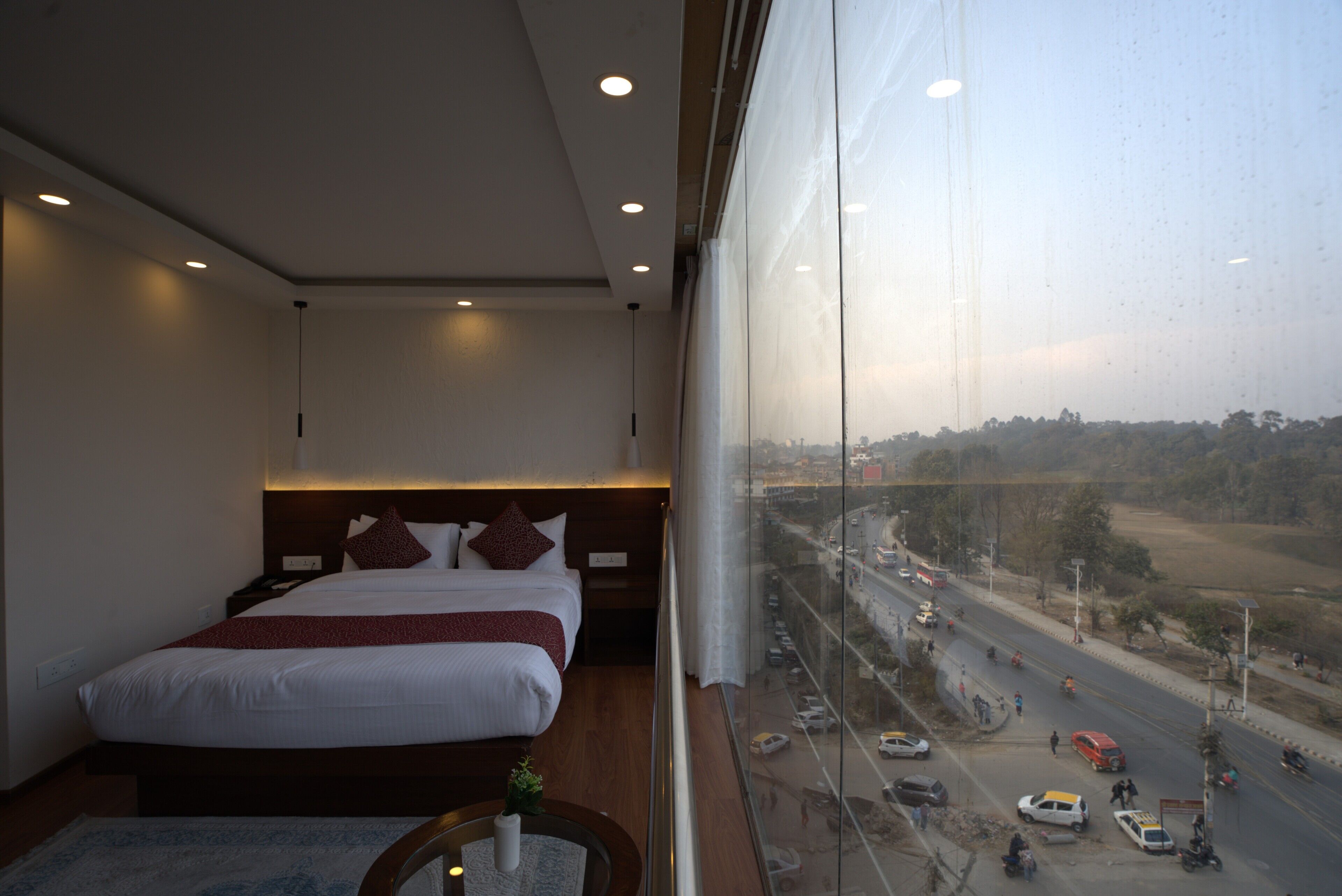 Ein Hotelzimmer mit einem großen Bett, einem Nachttisch und Blick auf eine belebte Straße durch ein großes Fenster.