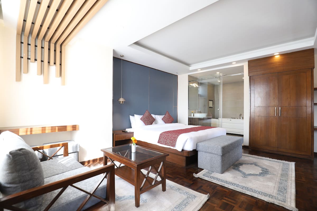 A modern hotel room with a bed, a desk, a chair, a wardrobe, and a bathroom visible through a glass door.