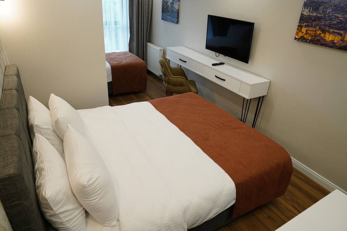 A hotel room with a bed, a desk with a TV, a chair, and a window with curtains.