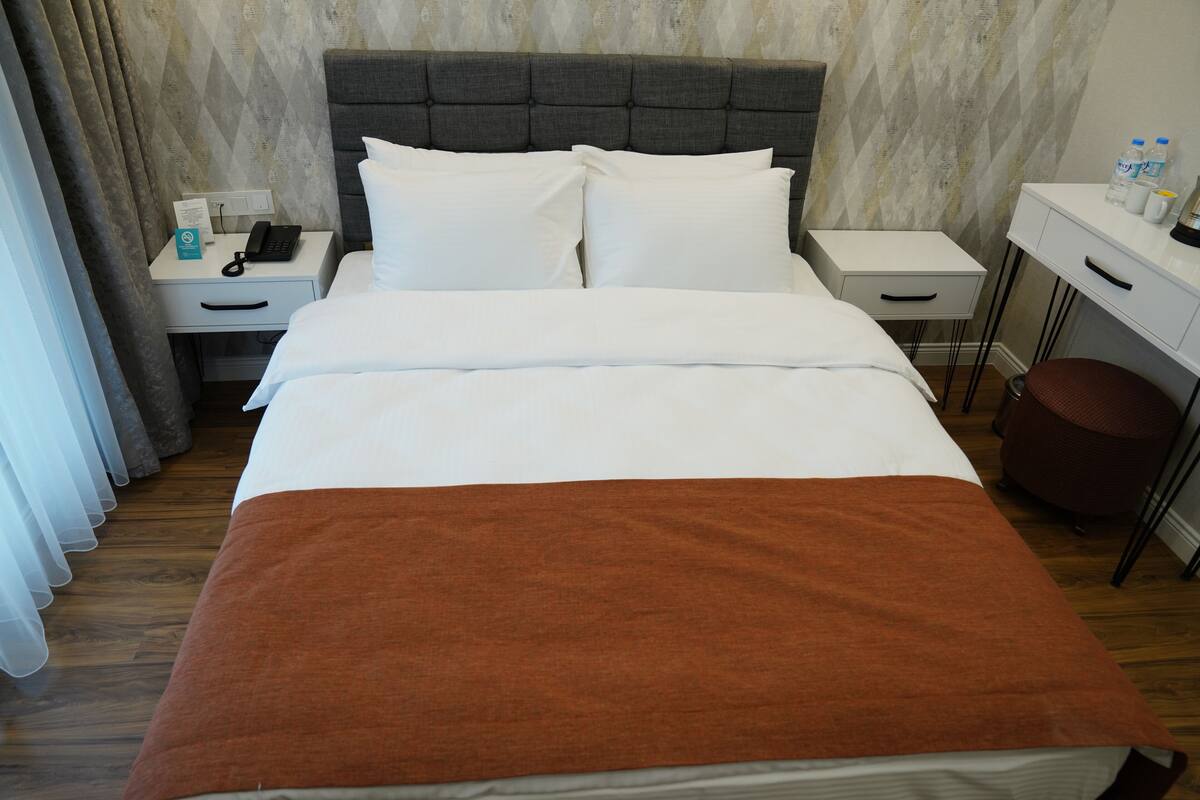 A neatly made bed with white linens and a brown throw blanket, a bedside table with a phone and a notepad, a small desk with a lamp, and a chair.