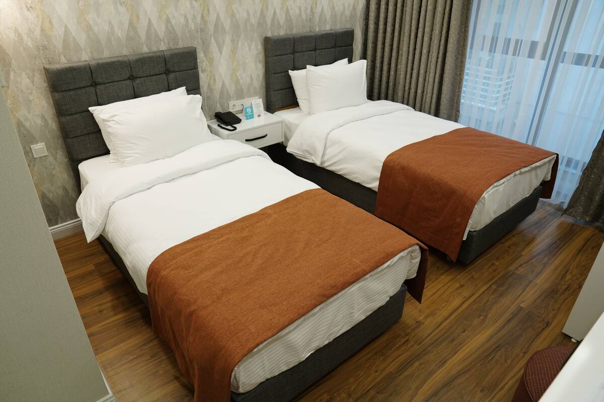 A hotel room with two beds, wooden flooring, a grey headboard, white bedding, and a nightstand with a phone and a card.