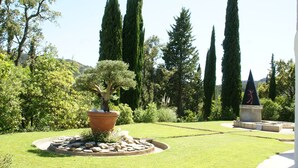 A well-maintained garden with a variety of trees, a central ornamental tree in a pot, and a stone monument. - Beautiful apartment near Brittany (Mandelieu-la-Napoule)