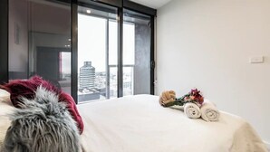A hotel room with a large sliding glass door to a city view, a bed with a fur throw, and a small table with towels and flowers.