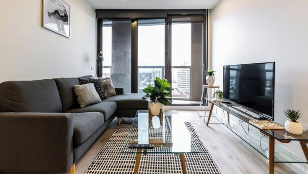 A modern living room with a sectional sofa, a glass coffee table, a flat-screen TV, and a large window offering a city view.