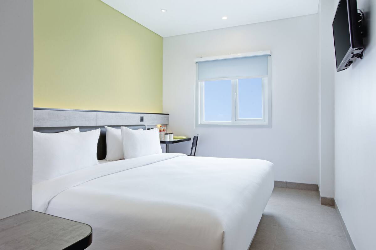 A hotel room with a bed, a small table, a chair, a window, and a TV mounted on the wall.
