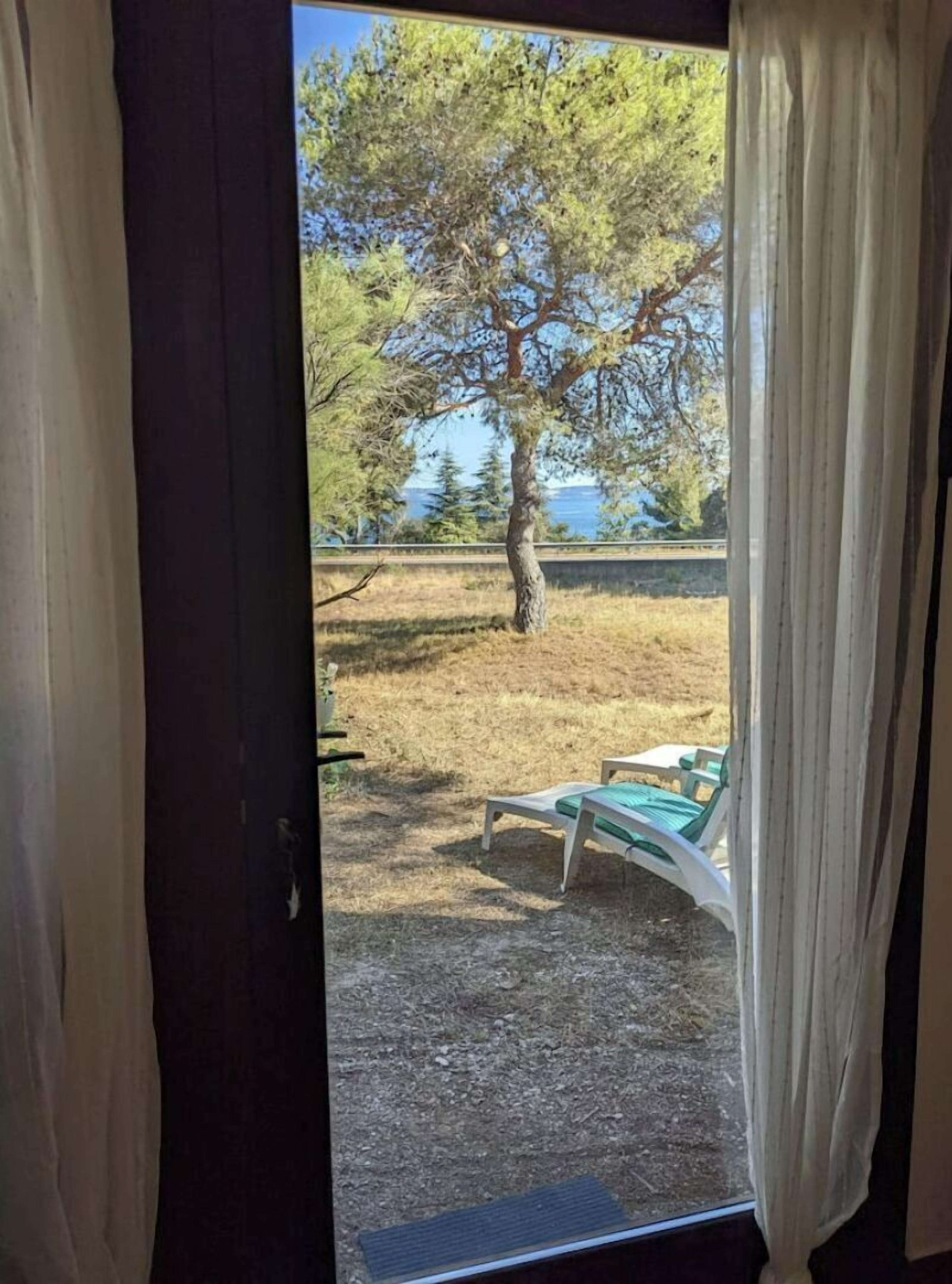 A room with a view of a tree and a body of water. There are two lounge chairs outside the window.