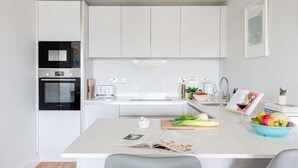 Une cuisine moderne avec des meubles blancs, un four encastré et un îlot central doté d’une planche à découper et de fruits.