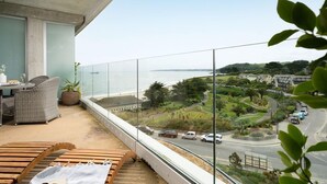 Un balcon avec vue sur la mer et une route. On y trouve une chaise en osier et un banc en bois.
