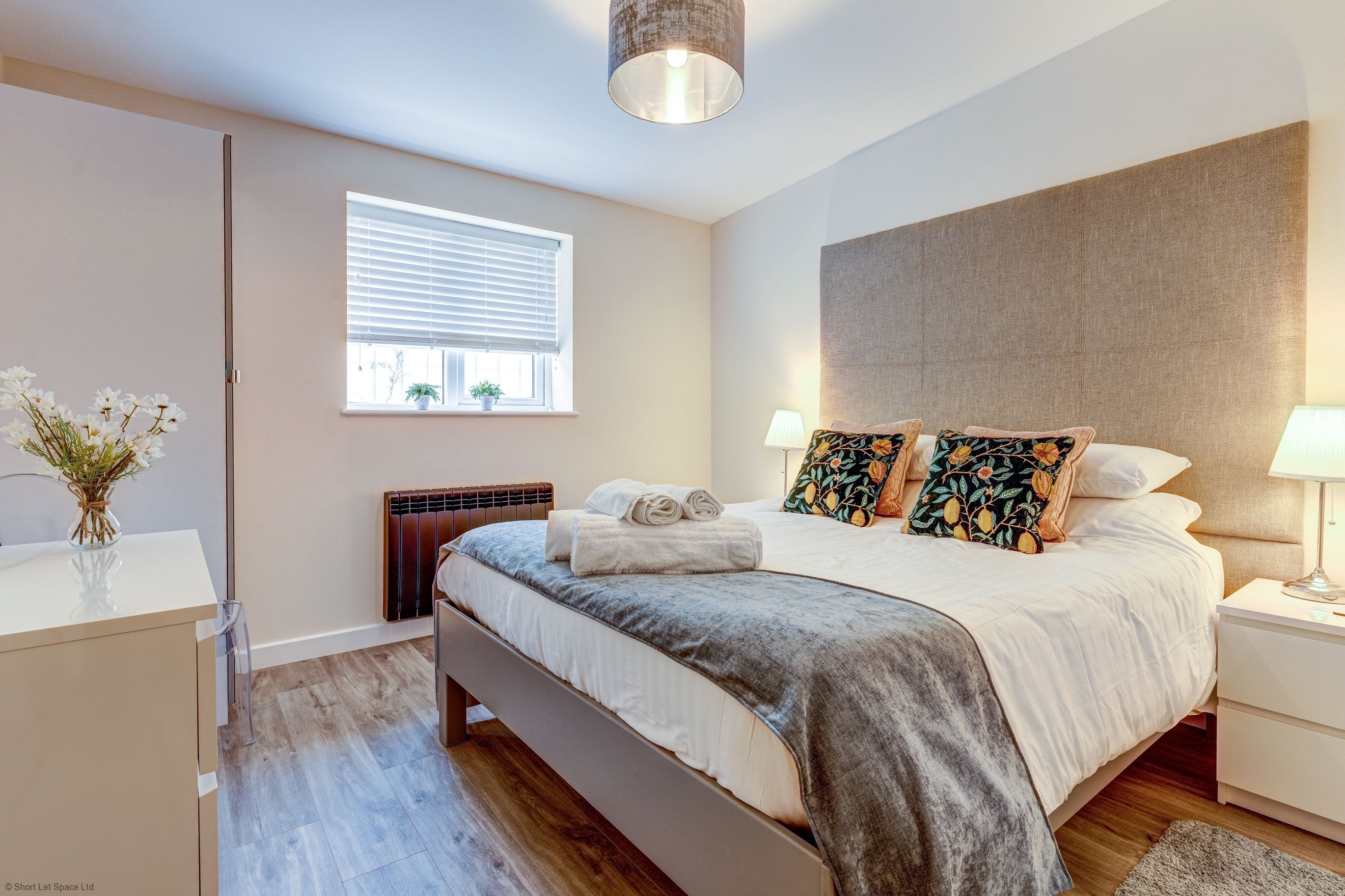 A modern bedroom with a large bed, bedside tables, a window with blinds, a radiator, and a vase with flowers.