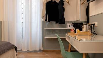 A modern hotel room with a bed, a desk with a coffee machine, a mirror, and a hanging clothes rack.