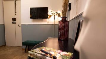 Een kamer met een houten vloer, een groene bank, een tafel met een notitieboekje en pennen met bloemendesign, een rode vaas met gedroogde planten en een flatscreen televisie aan de muur.